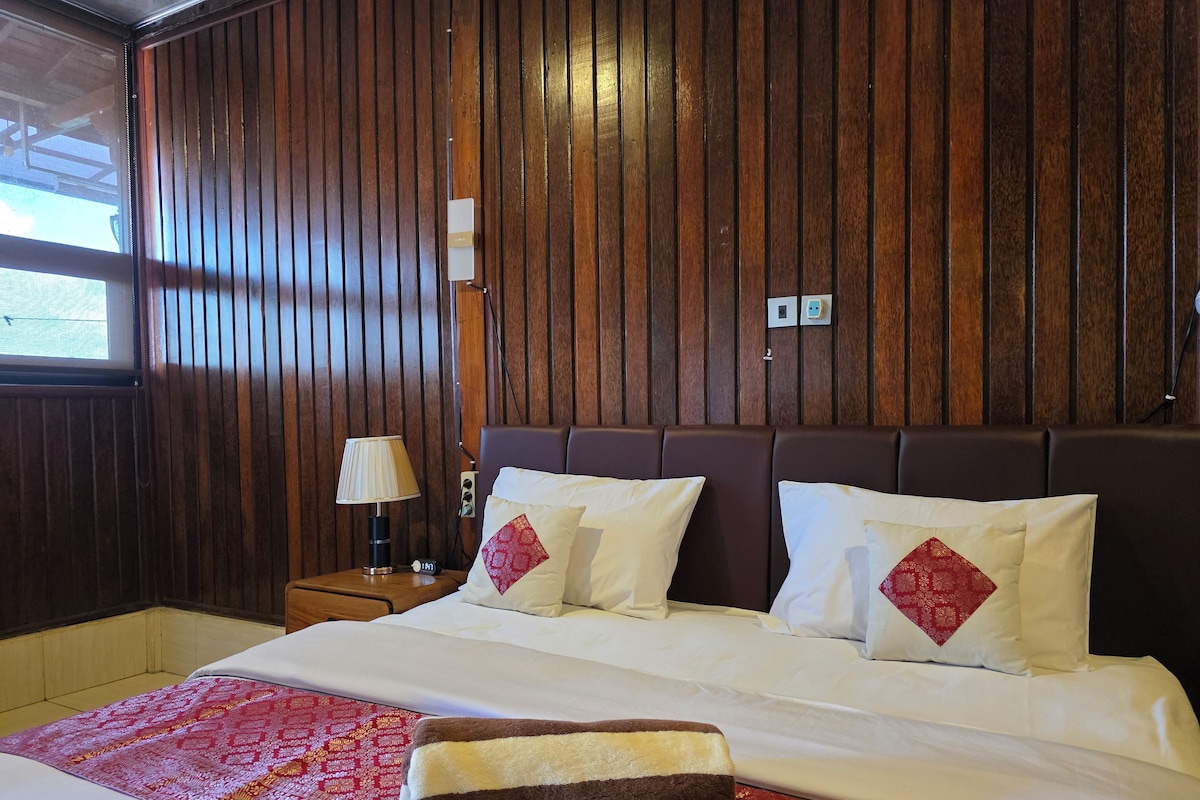 A hotel room with a wooden paneled wall, a bed with white bedding and red pillows, a nightstand with a lamp, and a red patterned bedspread.