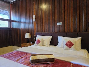 A hotel room with a wooden paneled wall, a bed with white bedding and red pillows, a nightstand with a lamp, and a red patterned bedspread. - Dahlia Moon Saba Beach (Blahbatuh)