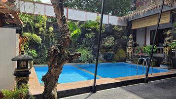 Ein Swimmingpool mit einem Baum im Vordergrund, einer Felswand mit Pflanzen und einer Statue rechts.