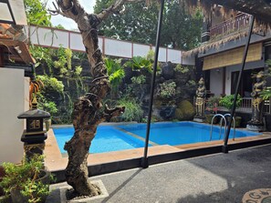 A swimming pool with a tree in the foreground, a rock wall with plants, and a statue on the right.