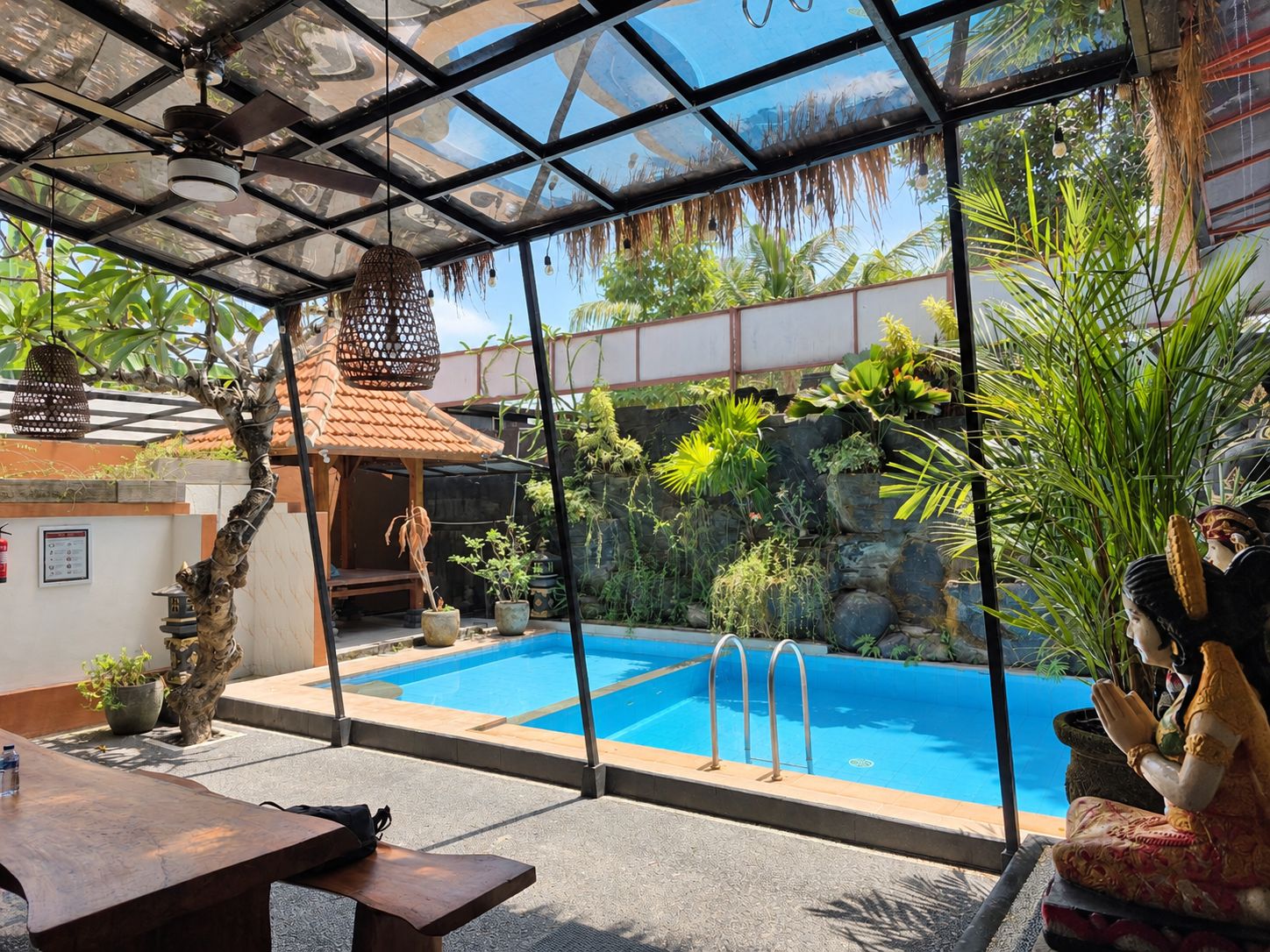 Área de piscina com teto de vidro, uma estátua e plantas tropicais.
