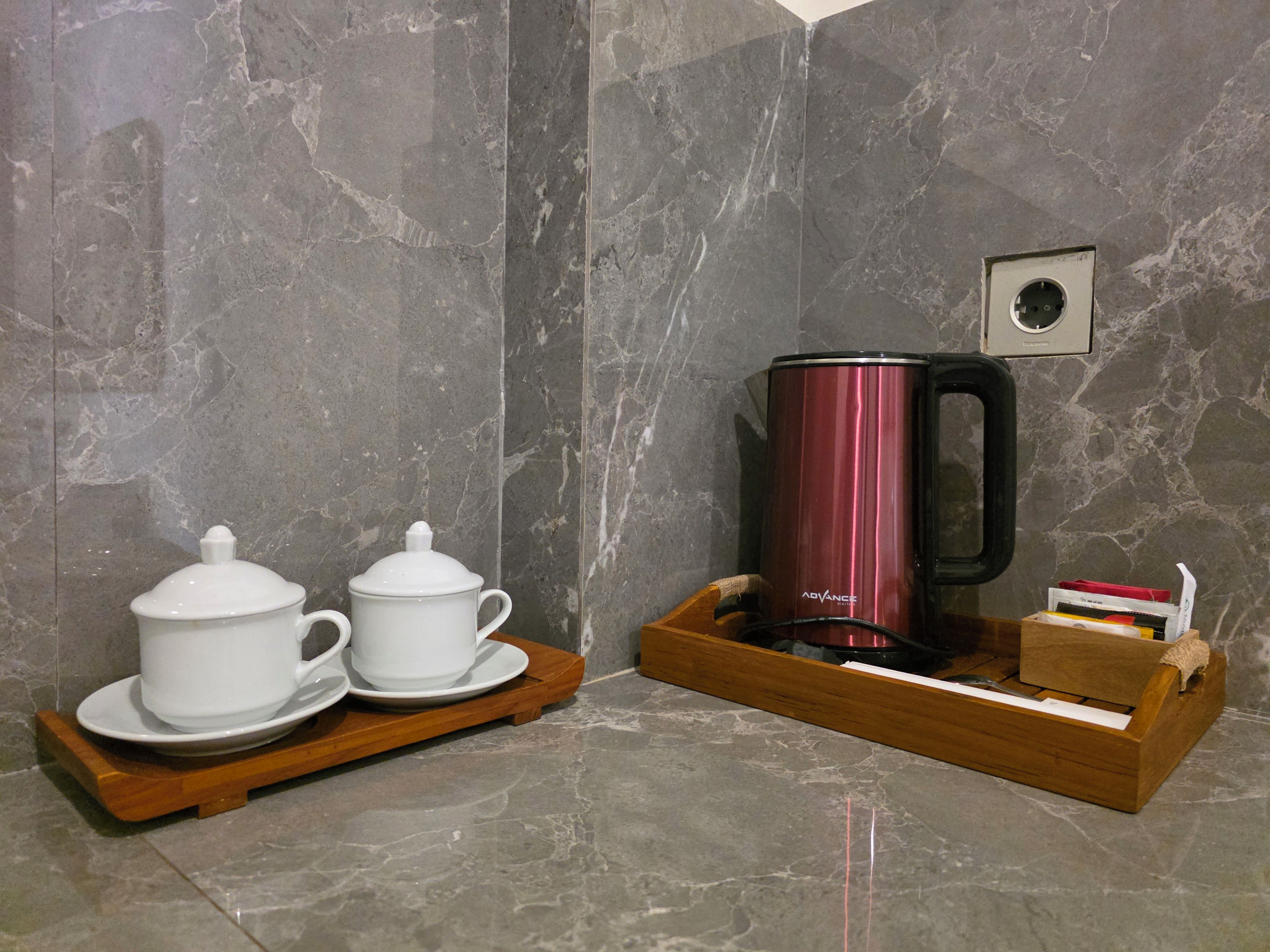 A marble wall with a coffee maker on a wooden tray, a white teapot, and a white cup on a matching tray.