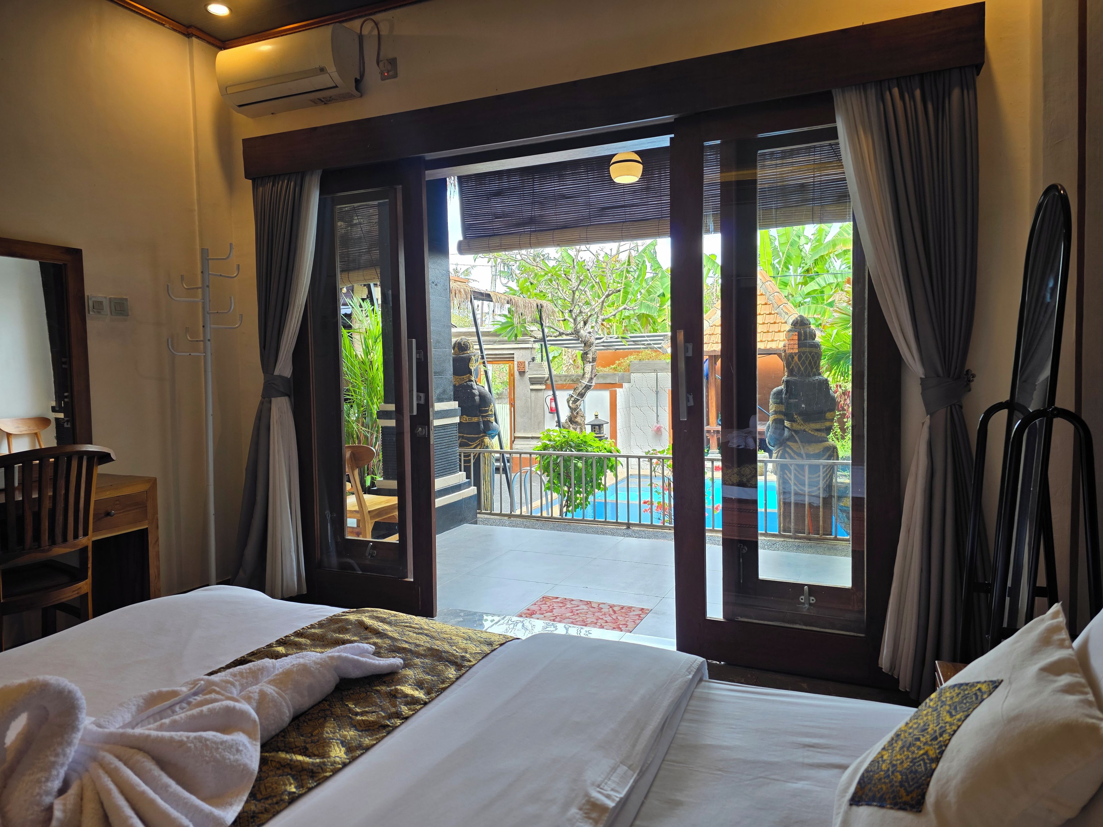 Quarto de hotel com cama, área de estar com poltrona e vista para área externa com estátua e plantas.