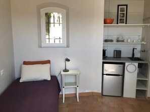 Ein gemütlicher Raum mit Bett, kleinem Tisch, Schreibtisch, Kühlschrank und Küchenzeile mit Regalen und Spüle.