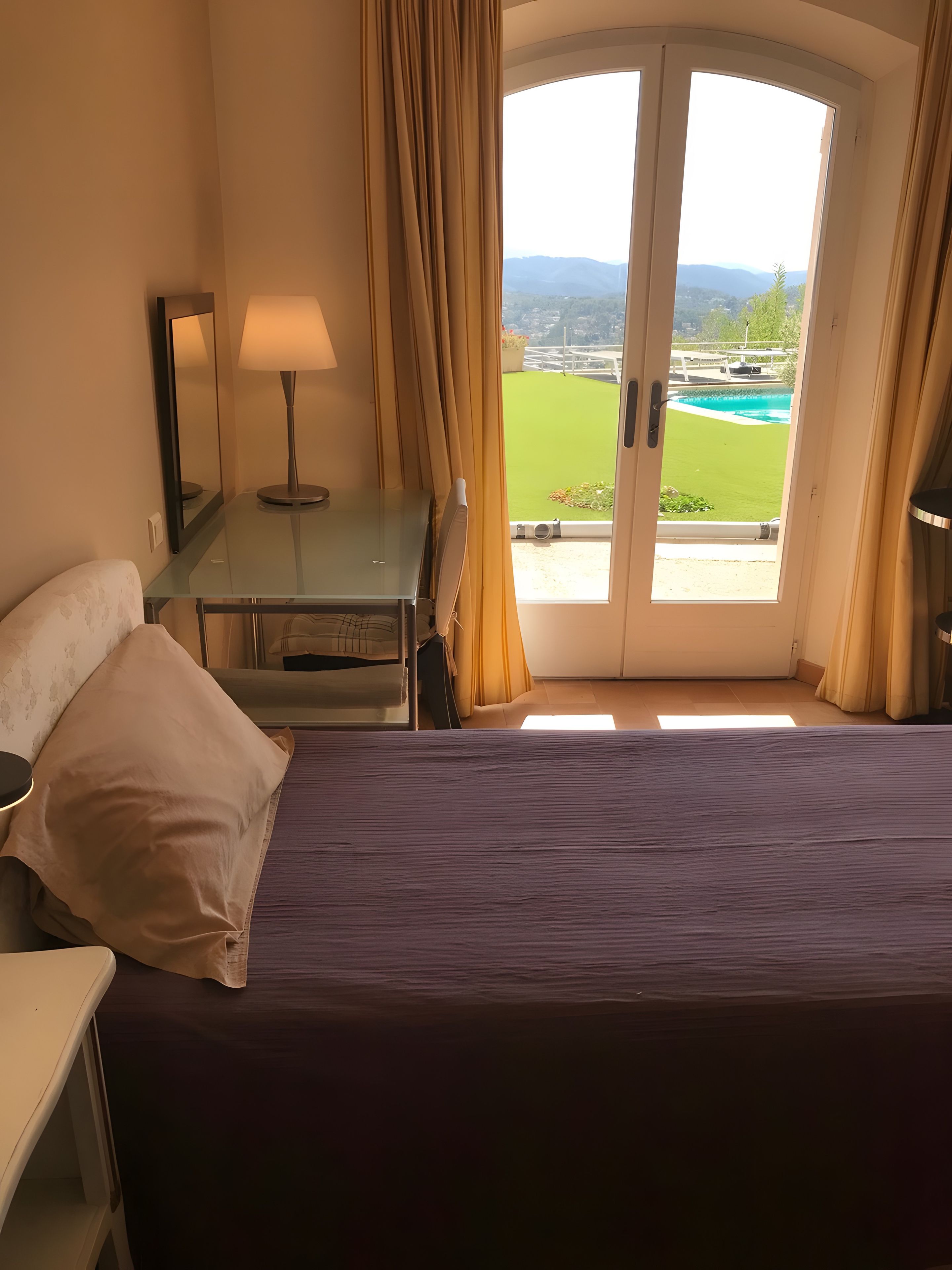 A hotel room with a bed, a desk, a lamp, and a view of a green lawn and pool through a glass door.