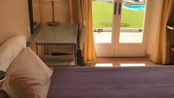A hotel room with a bed, a desk, a lamp, and a view of a green lawn and pool through a glass door.
