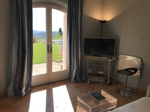 Ein Zimmer mit Fernseher, einem Sessel, einem Glastisch mit einem Buch und Blick auf eine grüne Landschaft durch eine Glastür.