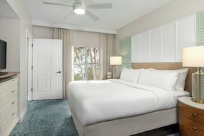 Una habitación de hotel con cama, lámparas de noche, ventilador de techo, ventana con cortinas y una puerta.