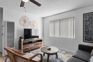 Ein Wohnzimmer mit einem Fernseher auf einem Holzboden, einem Deckenventilator, einem Fenster mit Jalousien und einem gemusterten Teppich.