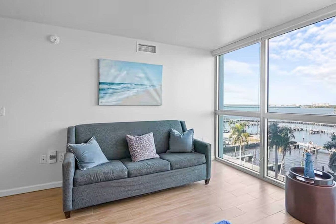 A modern living room with a grey sofa, a painting of a beach, and a large window overlooking a marina.