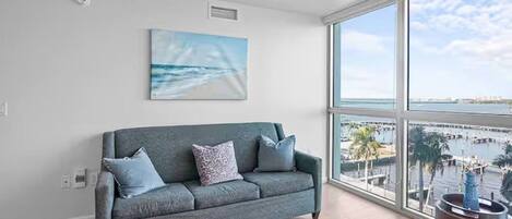 A modern living room with a grey sofa, a painting of a beach, and a large window overlooking a marina.