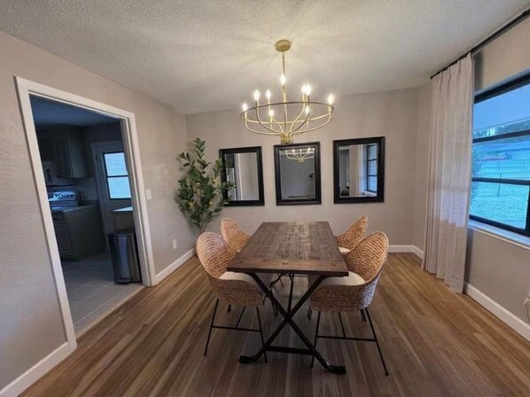 A dining area with a wooden table and wicker chairs, a chandelier, and a view of the outdoors.