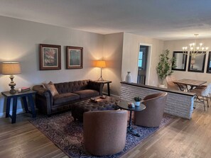 A living room with a brown leather sofa, a round coffee table, and two armchairs. There is a rug on the floor, a lamp on a side table, and framed artwork on the wall.