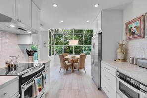 Una cocina moderna con armarios blancos, un comedor con sillas de mimbre y un amplio ventanal con vistas a la vegetación.