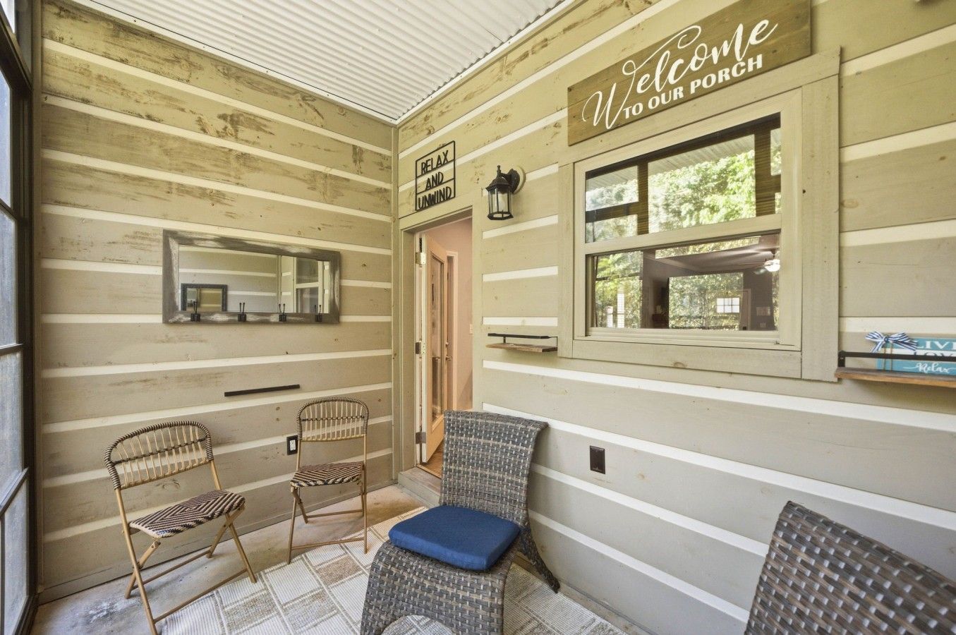 A wooden porch with a 'Welcome to our porch' sign, two wicker chairs, a bench with a cushion, a mirror, and a window with a view of greenery.