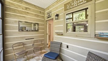 A wooden porch with a 'Welcome to our porch' sign, two wicker chairs, a bench with a cushion, a mirror, and a window with a view of greenery.