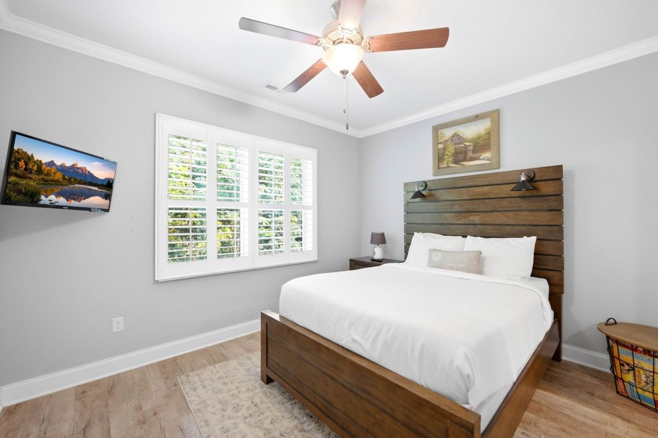 Une chambre à coucher avec un lit, un ventilateur de plafond, une fenêtre avec des stores, une table de chevet, une lampe et un tableau au mur.