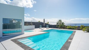 Una piscina exterior moderna con agua azul cristalina, rodeada por un muro de piedra y un edificio blanco con amplias ventanas.