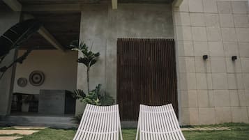 A modern building exterior with a concrete facade, a wooden door, and two white lounge chairs in the foreground.