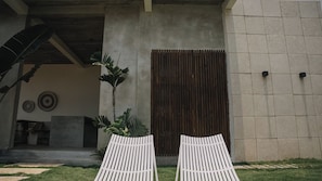 A modern building exterior with a concrete facade, a wooden door, and two white lounge chairs in the foreground.