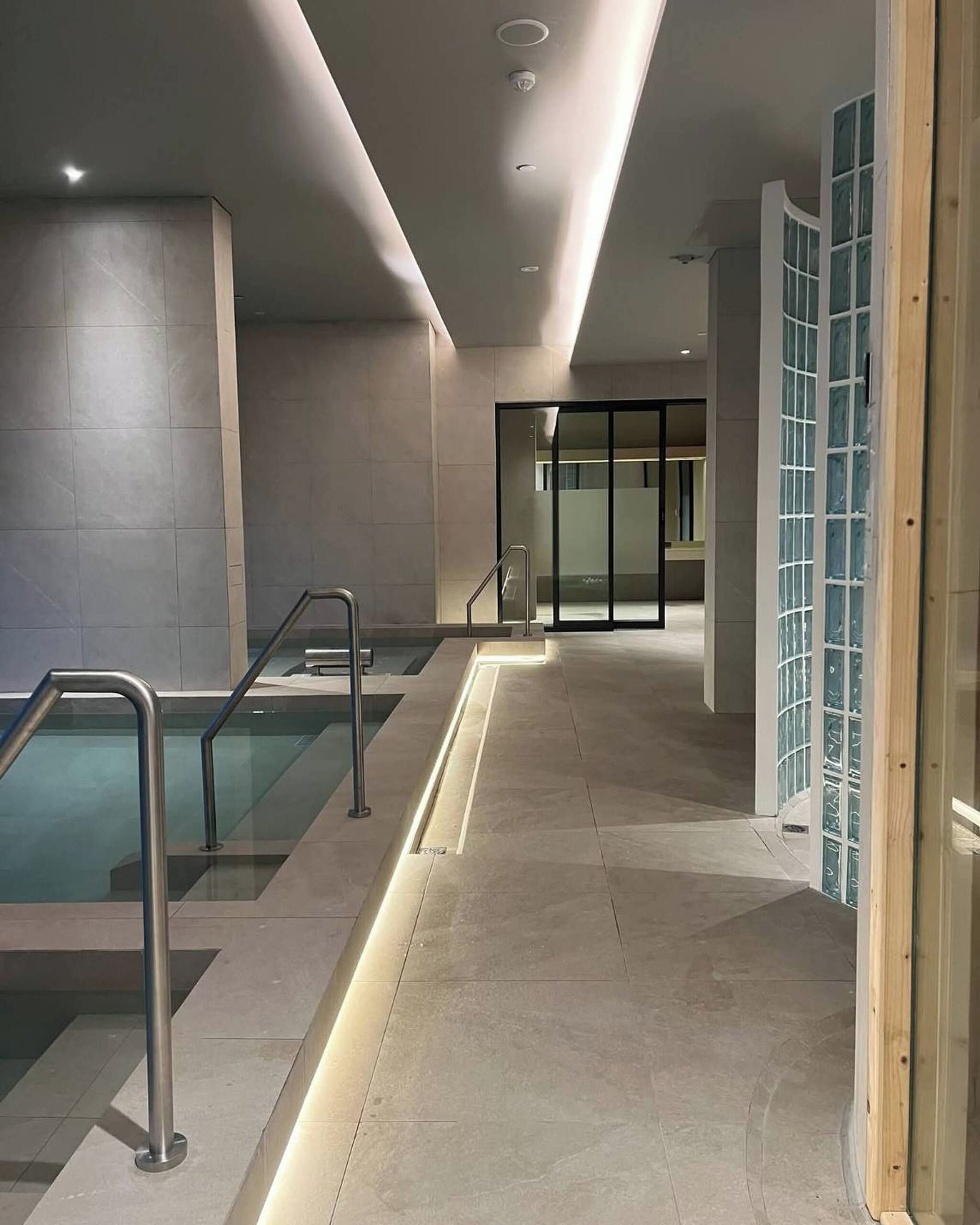 Área de piscina coberta moderna, com guarda-corpos de vidro transparente, piso ladrilhado e porta de vidro que dá para outro espaço.