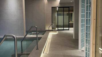 Área de piscina coberta moderna, com guarda-corpos de vidro transparente, piso ladrilhado e porta de vidro que dá para outro espaço.