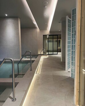 Área de piscina coberta moderna, com guarda-corpos de vidro transparente, piso ladrilhado e porta de vidro que dá para outro espaço.