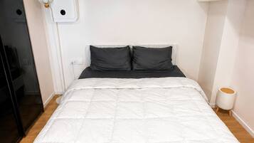 Quarto individual com uma cama de solteiro, colchão branco, cabeceira preta e uma pequena mesa de cabeceira com luminária.