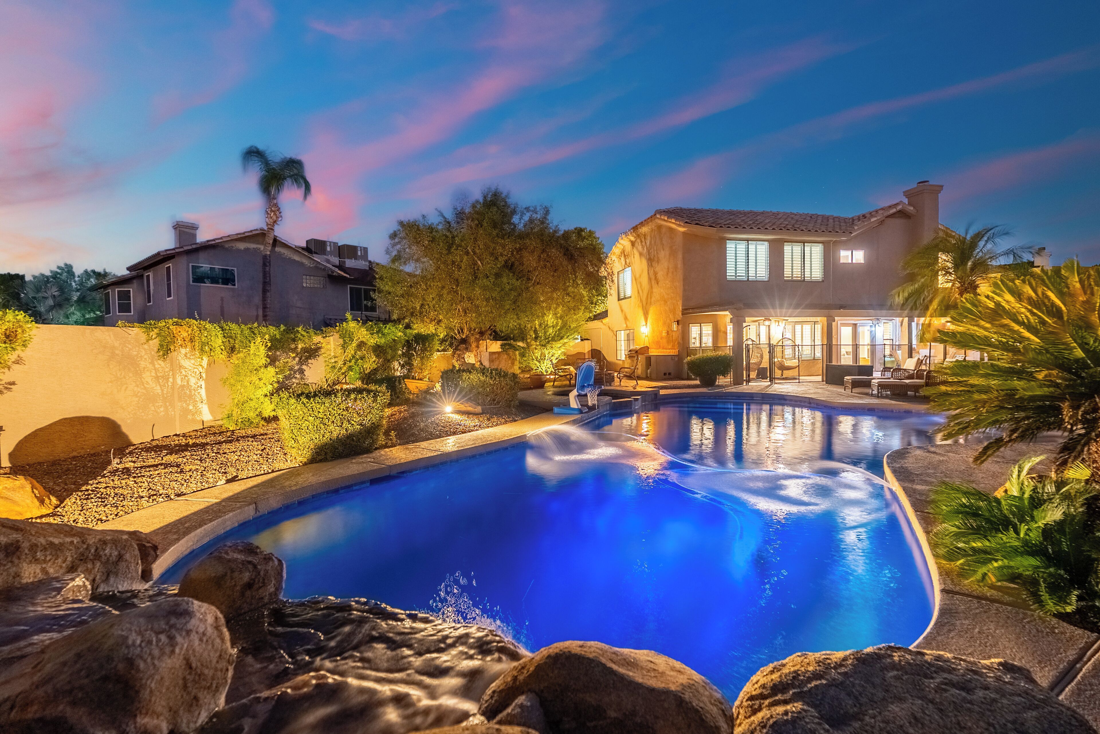A swimming pool with a waterfall feature, surrounded by rocks and plants, with a large house in the background.