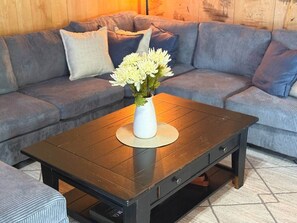 Ein Wohnzimmer mit einem dunkelholznen Couchtisch, einer weißen Vase mit Blumen und einem grauen Sofa mit Kissen.