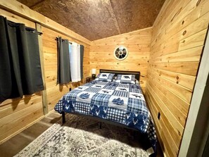 A wooden cabin bedroom with a bed, curtains, a window, and a wall-mounted fan. - Trail Access Getaway Near Lake Saint Helen (St. Helen)