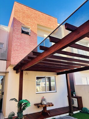 A residential building with a brick facade, a wooden pergola, a small table with a bag and a potted plant, and a well-maintained lawn.