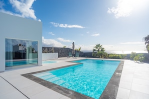 Piscina ao ar livre moderna com água azul cristalina, cercada por um deck de azulejos brancos e uma parede de pedra.