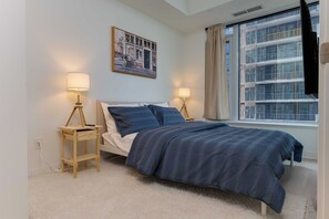 A bedroom with a bed, two bedside tables, a window with curtains, and a wall-mounted picture. - Stylish Yorkville 1BR Central & Convenient (Toronto)