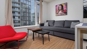 Ein modernes Wohnzimmer mit einem grauen Sofa, einem roten Sessel, einem kleinen Tisch und einem großen Fenster mit Blick auf ein Gebäude.