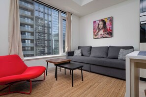 A modern living room with a grey sofa, a red chair, a small table, and a large window with a view of a building. - Stylish Yorkville 1BR Central & Convenient (Toronto)
