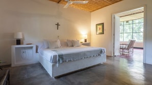 Una camera da letto con un letto, lampade sui comodini, una croce al muro e una porta che dà su uno spazio esterno.