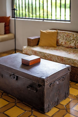 Une pièce avec une vieille valise posée sur un sol carrelé, un canapé recouvert d’un tissu à motifs floraux et une fenêtre donnant sur un paysage verdoyant.