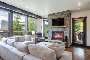 A modern living room with a stone fireplace, large windows, and a leather sofa. - Pet Friendly Mountain Retreat | Private Hot Tub | Private Firepit | Direct Trail Access (Silverthorne)