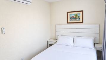 A hotel room with a bed, a nightstand, a bedside table, an air conditioning unit, a power outlet, and a framed picture on the wall.