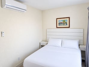 A hotel room with a bed, a nightstand, a bedside table, an air conditioning unit, a power outlet, and a framed picture on the wall.