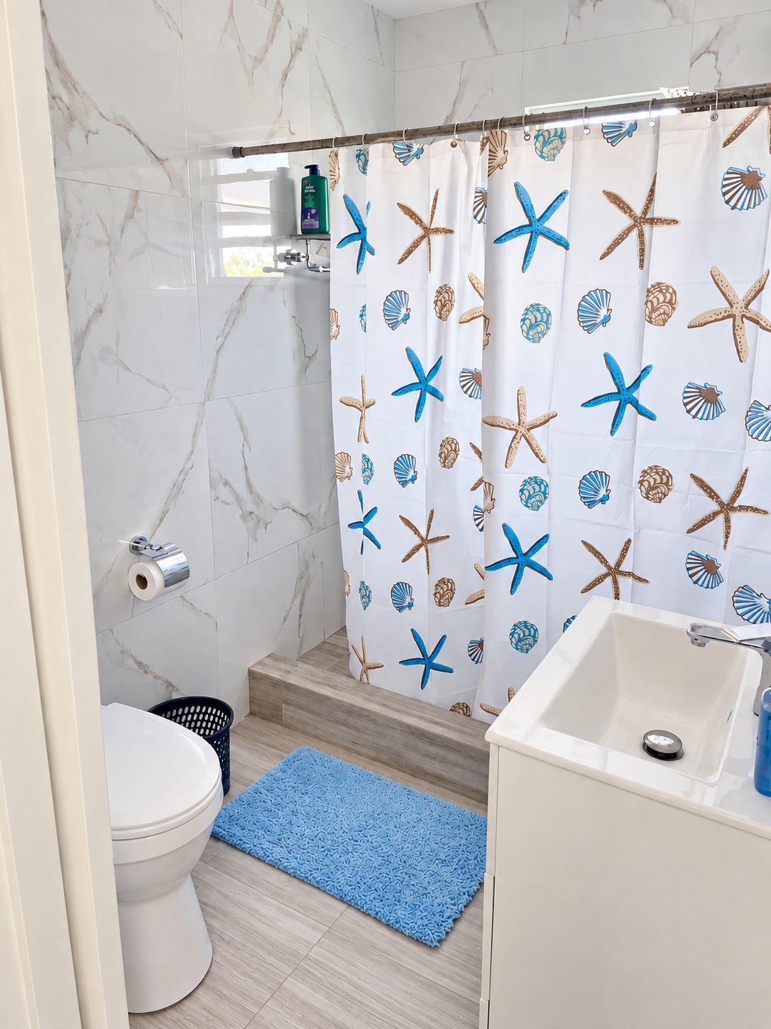 Un baño con pared de mármol blanco, cortina de ducha con estrellas de mar y conchas marinas, lavamanos blanco y felpudo de baño azul.