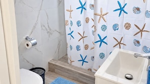 Ein Badezimmer mit weißer Marmorwand, einer Duschvorhang mit Seesternen und Muscheln, einem weißen Waschbecken und einer blauen Badmatte.