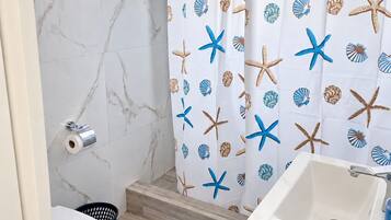 Un baño con pared de mármol blanco, cortina de ducha con estrellas de mar y conchas marinas, lavamanos blanco y felpudo de baño azul.