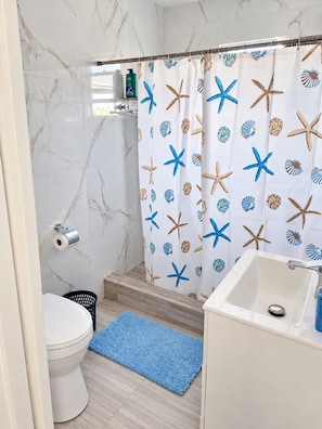 Een badkamer met een witte marmeren wand, een douchegordijn met zeesterren en schelpen, een wit wastafeltje en een blauwe badmat.