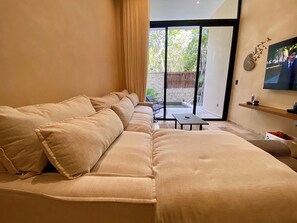 A living room with a beige sofa, a small table, and a TV mounted on the wall. - Super High Ceilings 2 BR, 2.5 Bath Apt. with plunge pool and terrace. (Tulum)