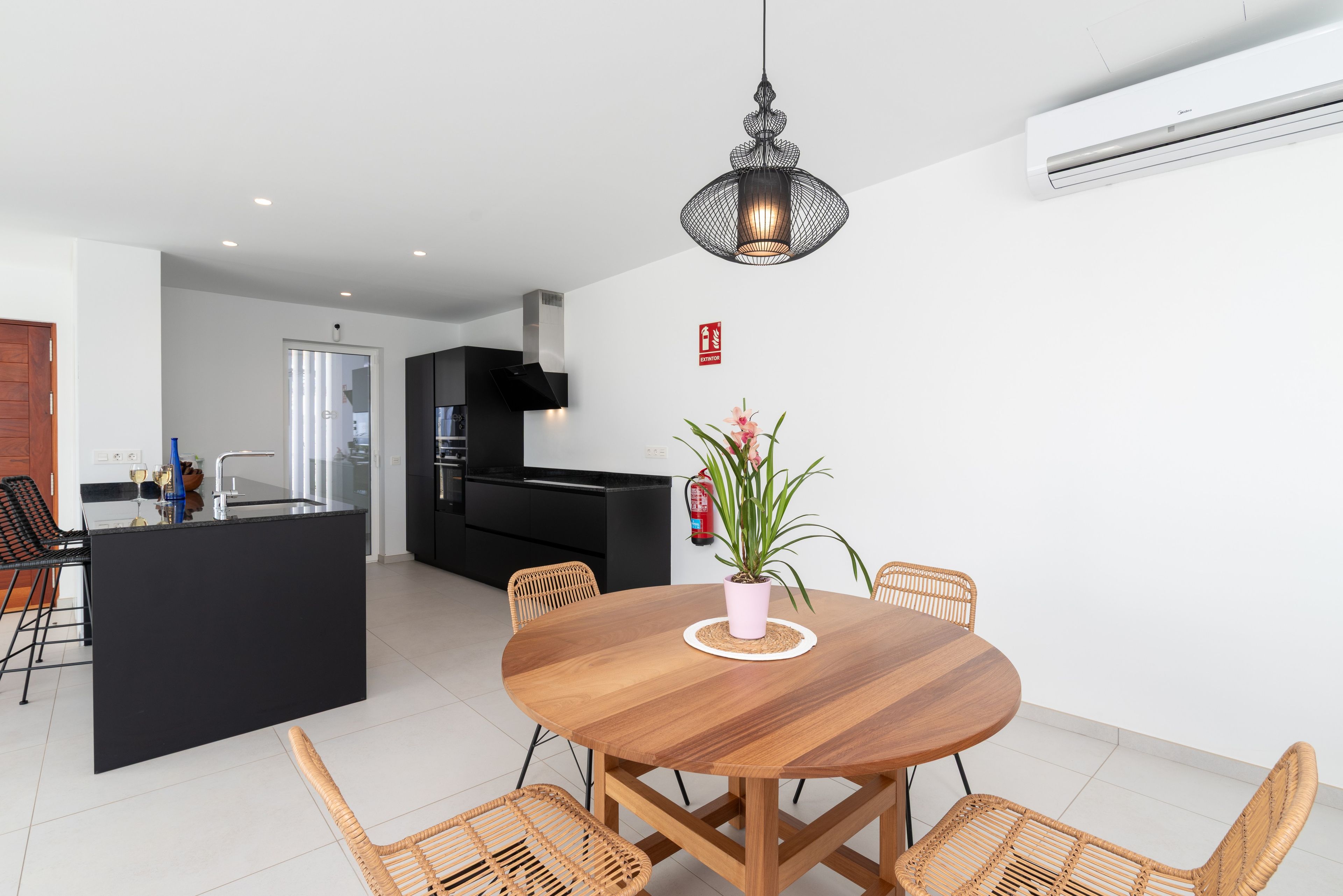 Una cocina moderna con armarios negros, una isla central y una zona de comedor con una mesa redonda de madera y sillas de mimbre.