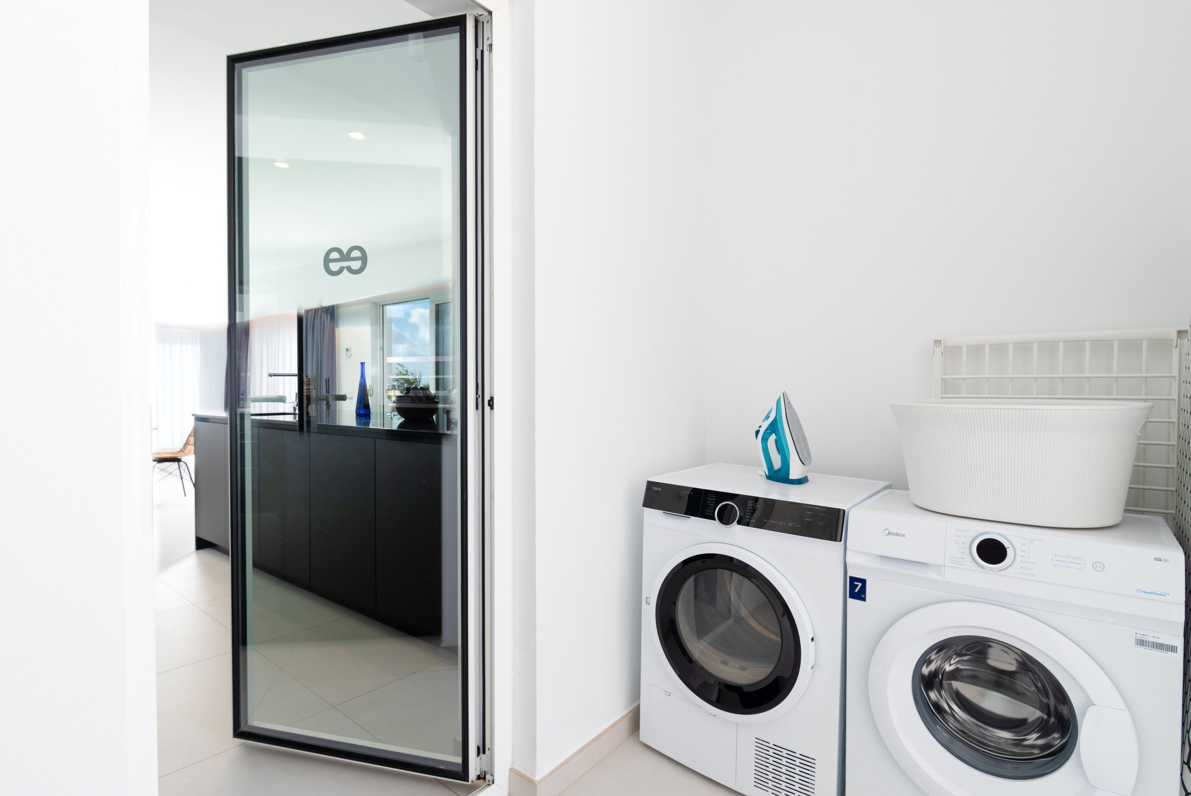 A modern laundry room with a front-loading washing machine and dryer, a glass door leading to a kitchen area, and a white tiled wall.