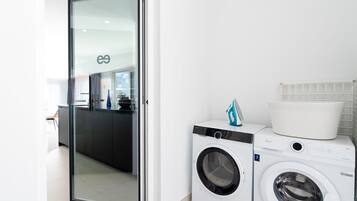 A modern laundry room with a front-loading washing machine and dryer, a glass door leading to a kitchen area, and a white tiled wall.