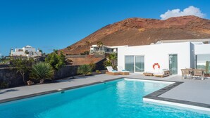 Uma área de piscina moderna com piscina azul cristalina, um edifício branco e uma montanha ao fundo.